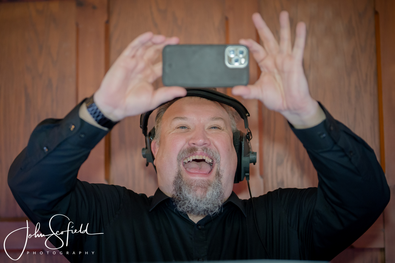 Ian smiling behind the booth, wearing headphones and holding a phone