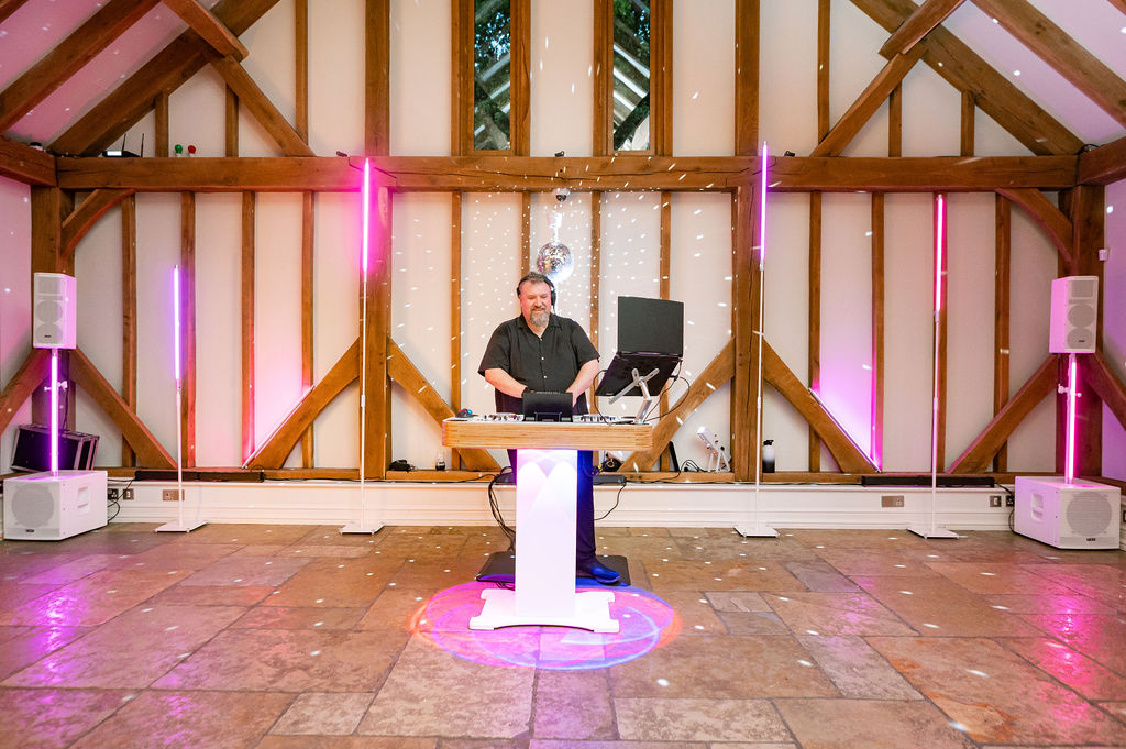 Ian at a white DJ setup with coordinated pink uplighting in a wedding venue